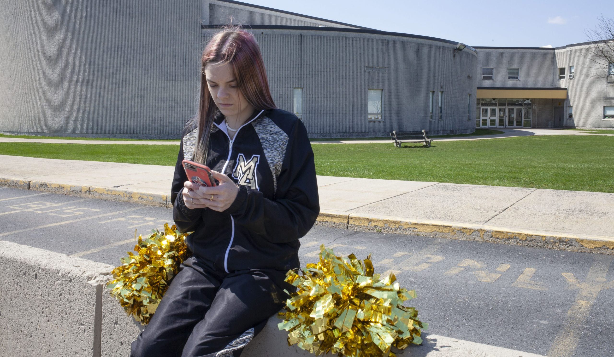 student holding phone and sitting in front of high school with cheerleading pom-poms beside her