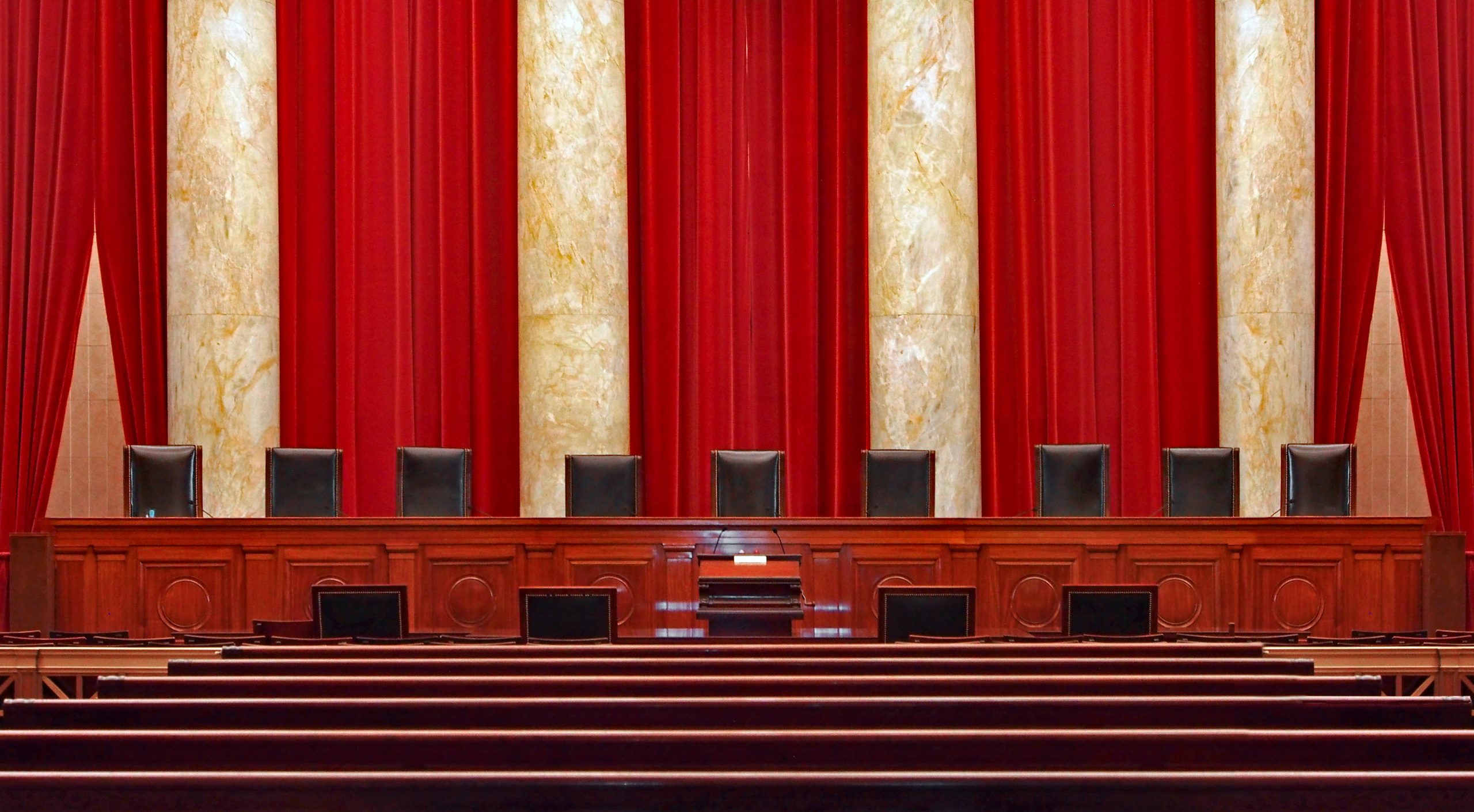 lectern in foreground in front of bench with nine empty chairs and tall red curtains in background