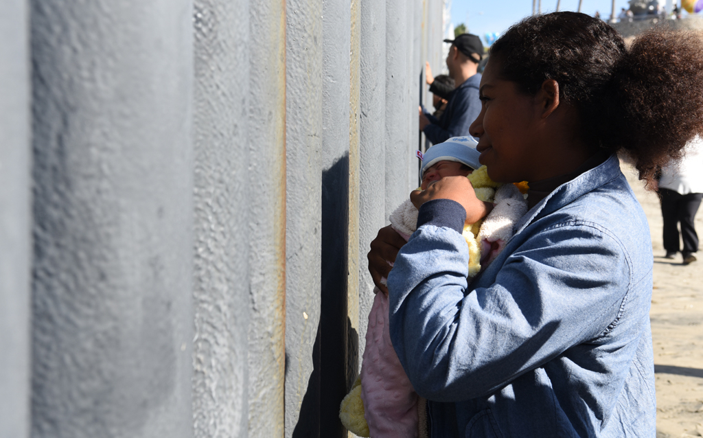 woman holding infant while standing a few inches from tall gray fence