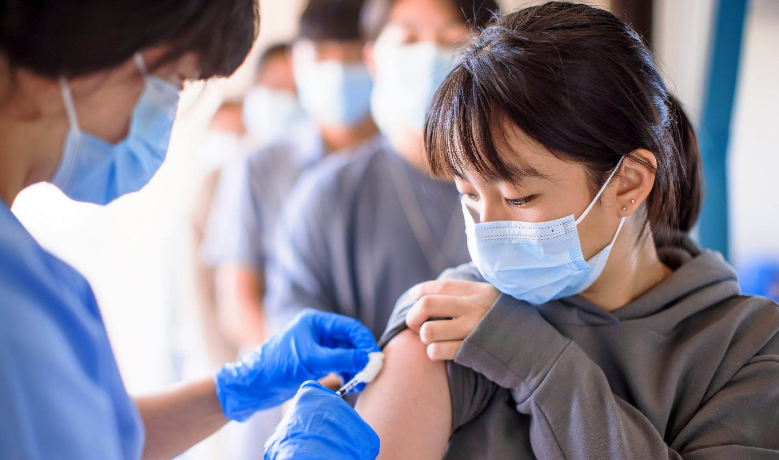 masked person wearing gloves administering vaccine to right arm of young person wearing mask