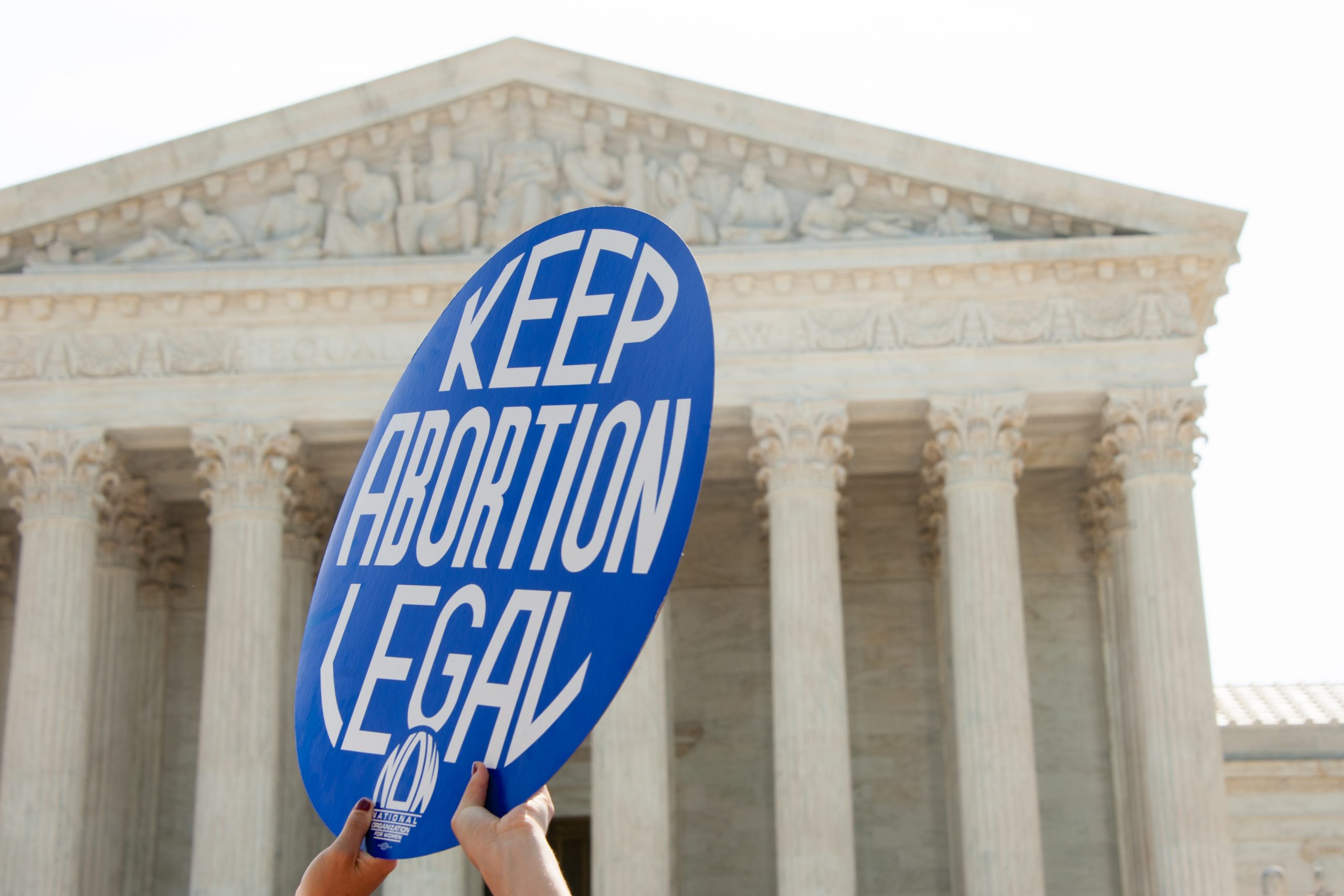 two hands holding round sign reading "Keep Abortion Legal" with Supreme Court building in background