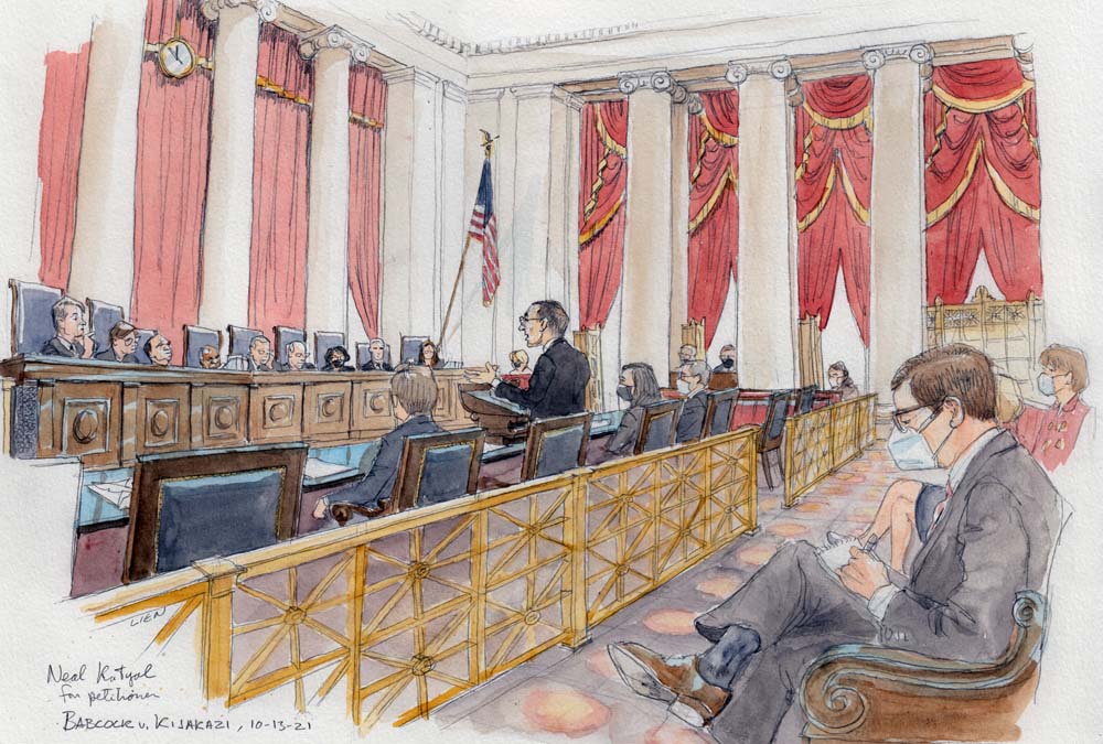 wide view of courtroom with nine justices sitting at bench, man standing at lectern, and several spectators wearing masks seated in gallery