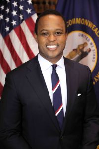 daniel cameron smiling in dark suit and striped tie with american flag in background