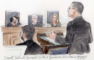 man at lectern with seated masked colleague in foreground and three justices in background