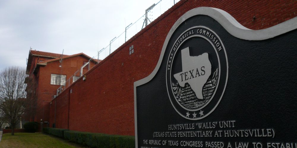 Texas historic marker in front of brick prison wall topped with barbed wire.