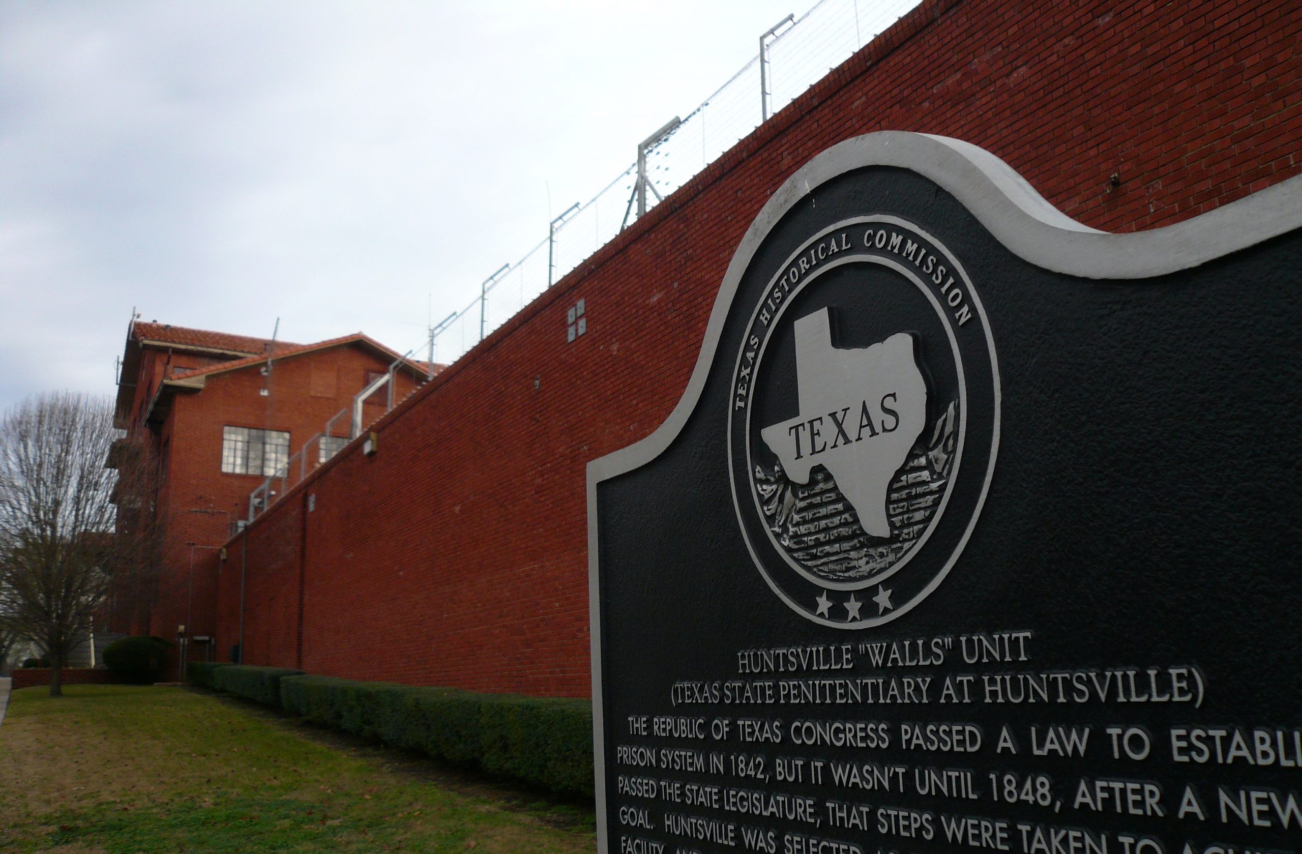 Texas historic marker in front of brick prison wall topped with barbed wire.