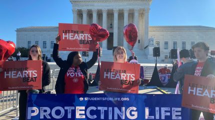 three women and one man hold signs reading "let their hearts beat" with supreme court building in background