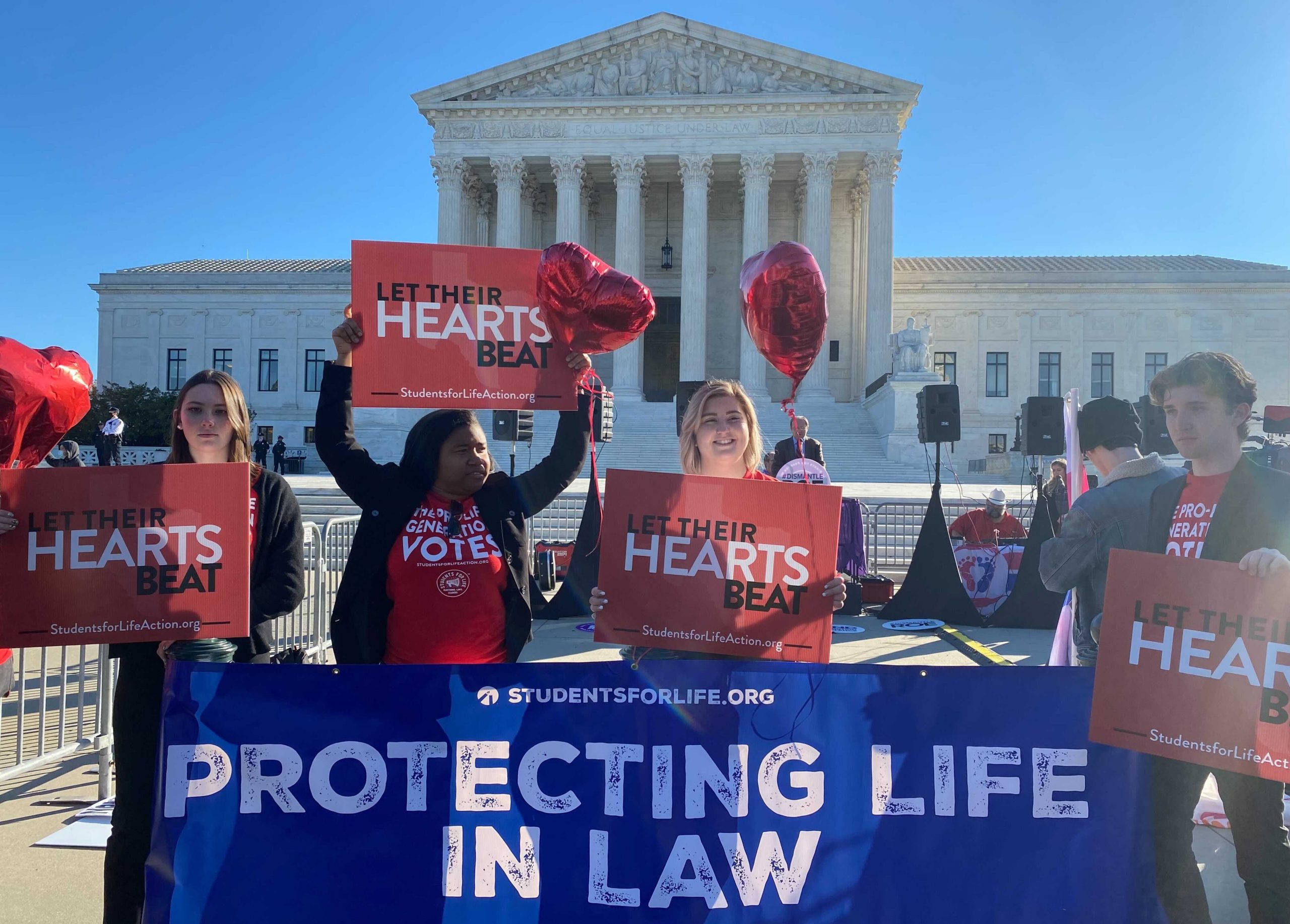 three women and one man hold signs reading "let their hearts beat" with supreme court building in background