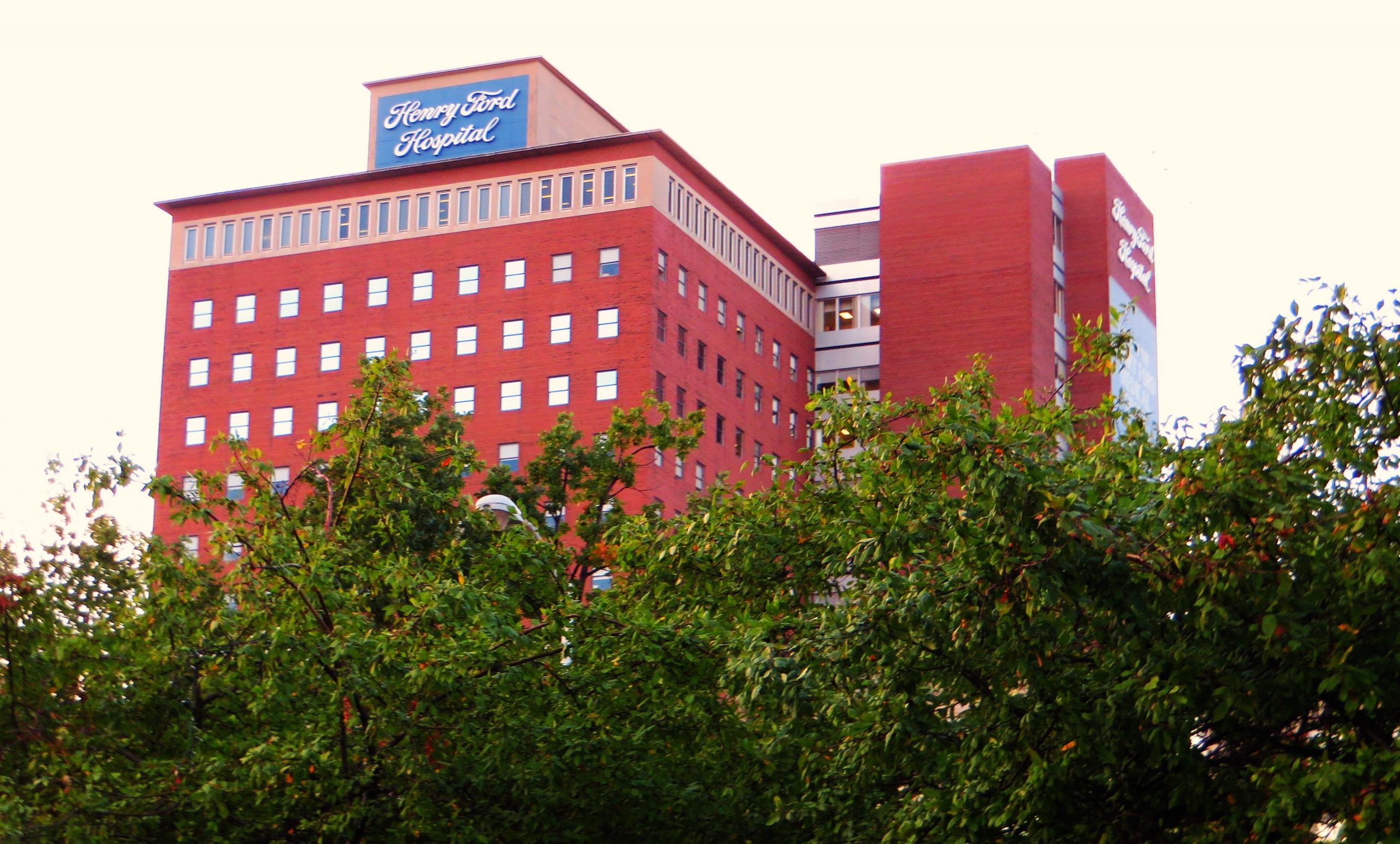 view from distance of large brick building with sign reading Henry Ford Hospital and tall trees in foreground