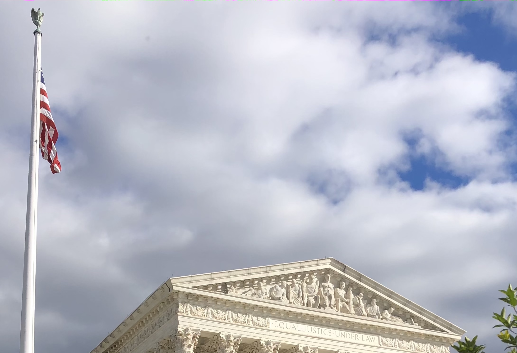 top of supreme court building with american flag on left against cloudy blue sky