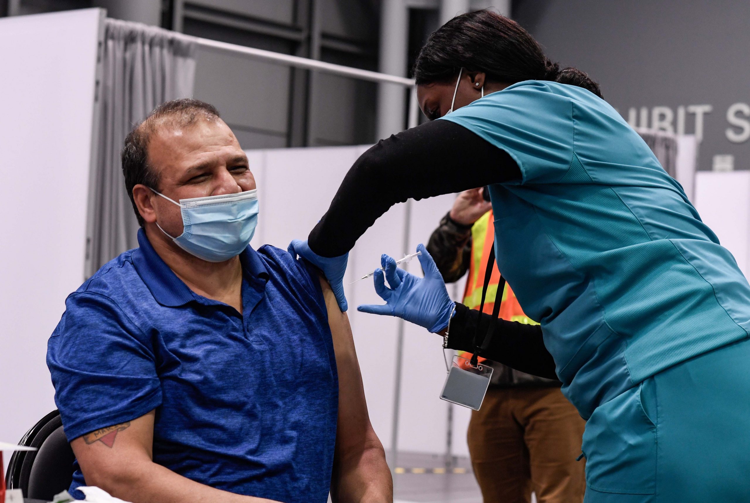 sitting man wearing mask receiving a vaccine in left arm from standing woman in scrubs