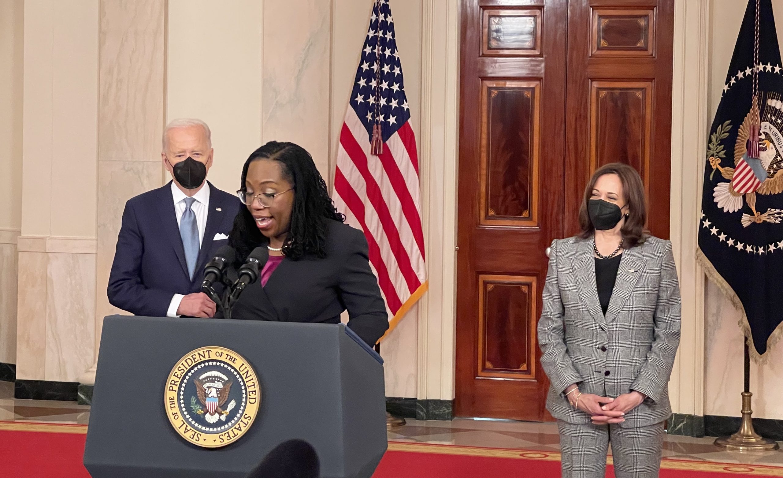 woman speaking at lectern with joe biden and kamala harris standing in background wearing masks