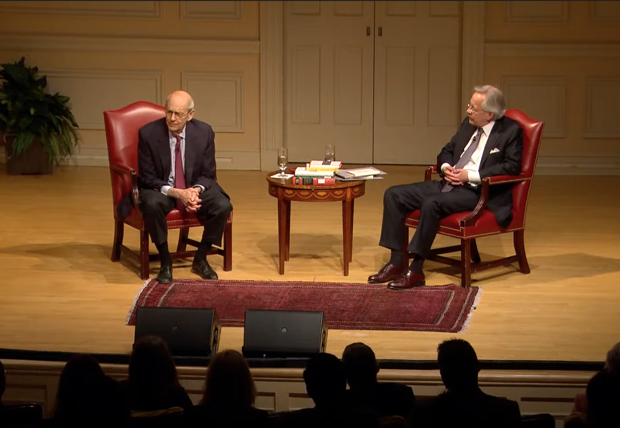 two men seated, speaking, on stage