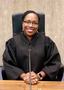 woman in black judicial robe sitting in front of microphone with hands folded together, smiling broadly