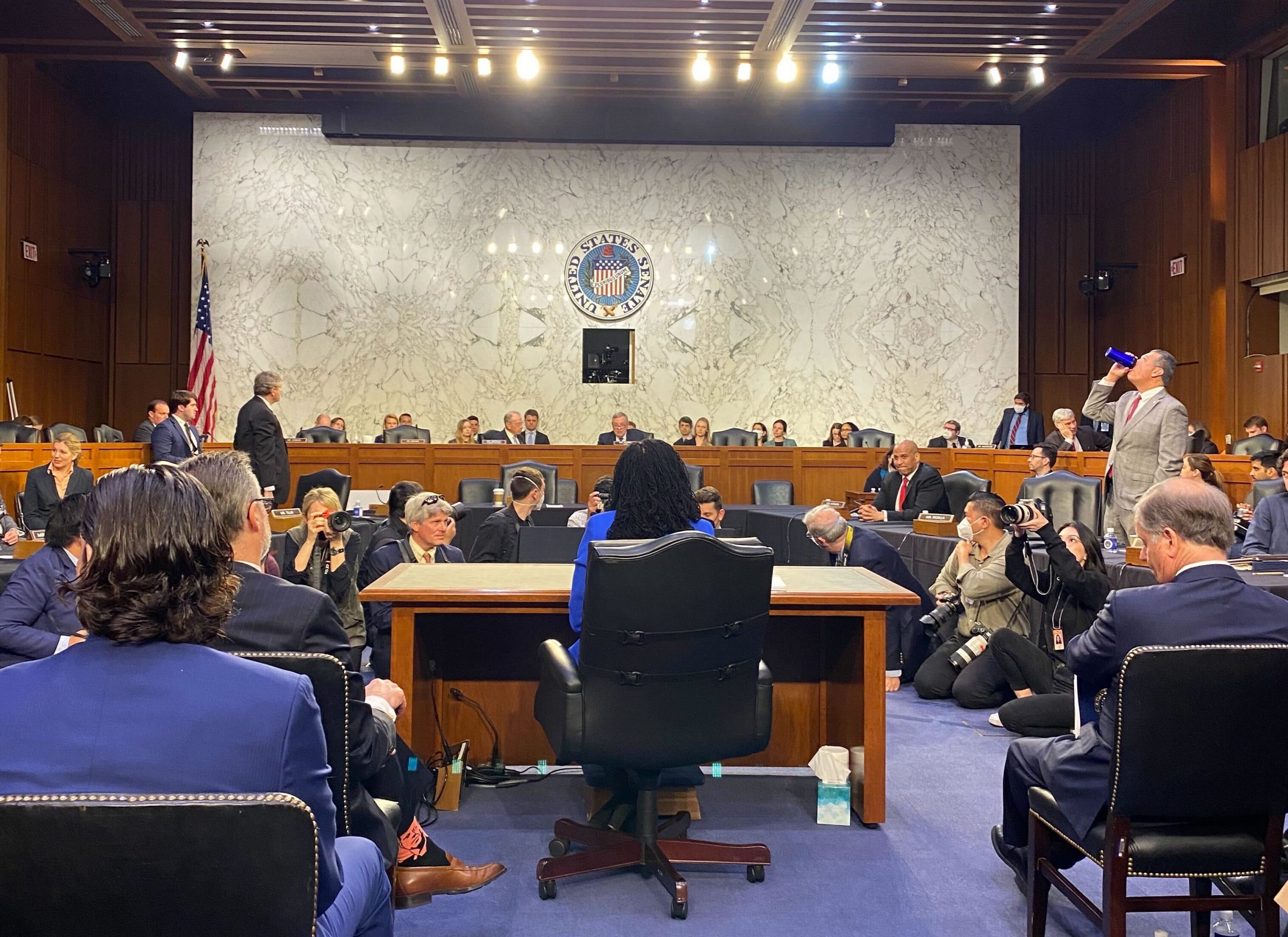 ketanji brown jackson sitting at desk, facing away from camera and toward crowd of photographers and senators sitting on dais in background