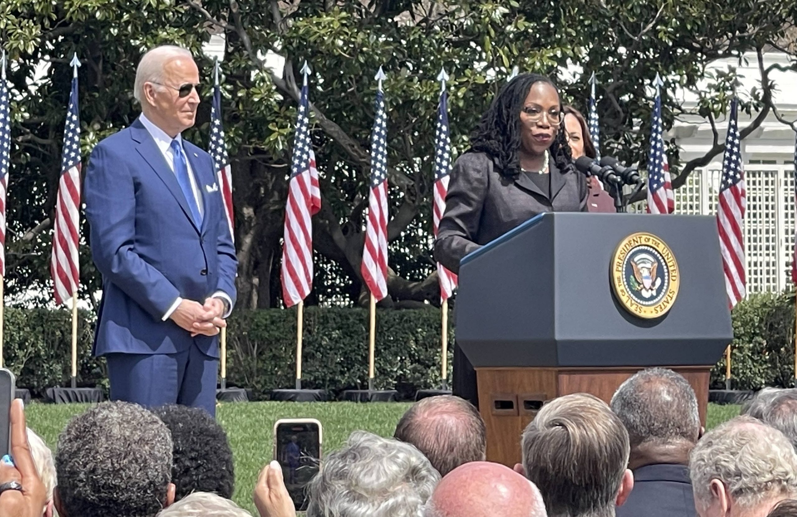 Ketanji Brown Jackson stands speaking at a lectern in front of a crowd outdoors. Joe Biden looks on with hands clasped in front of him.