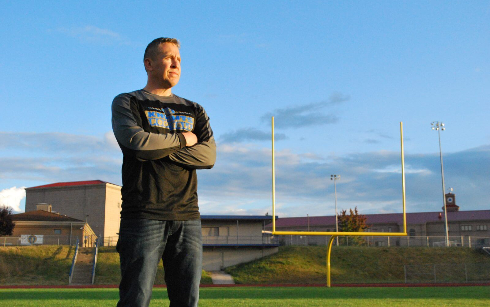 man standing alone on football field with arms crossed. field goal uprights are seen in the background.