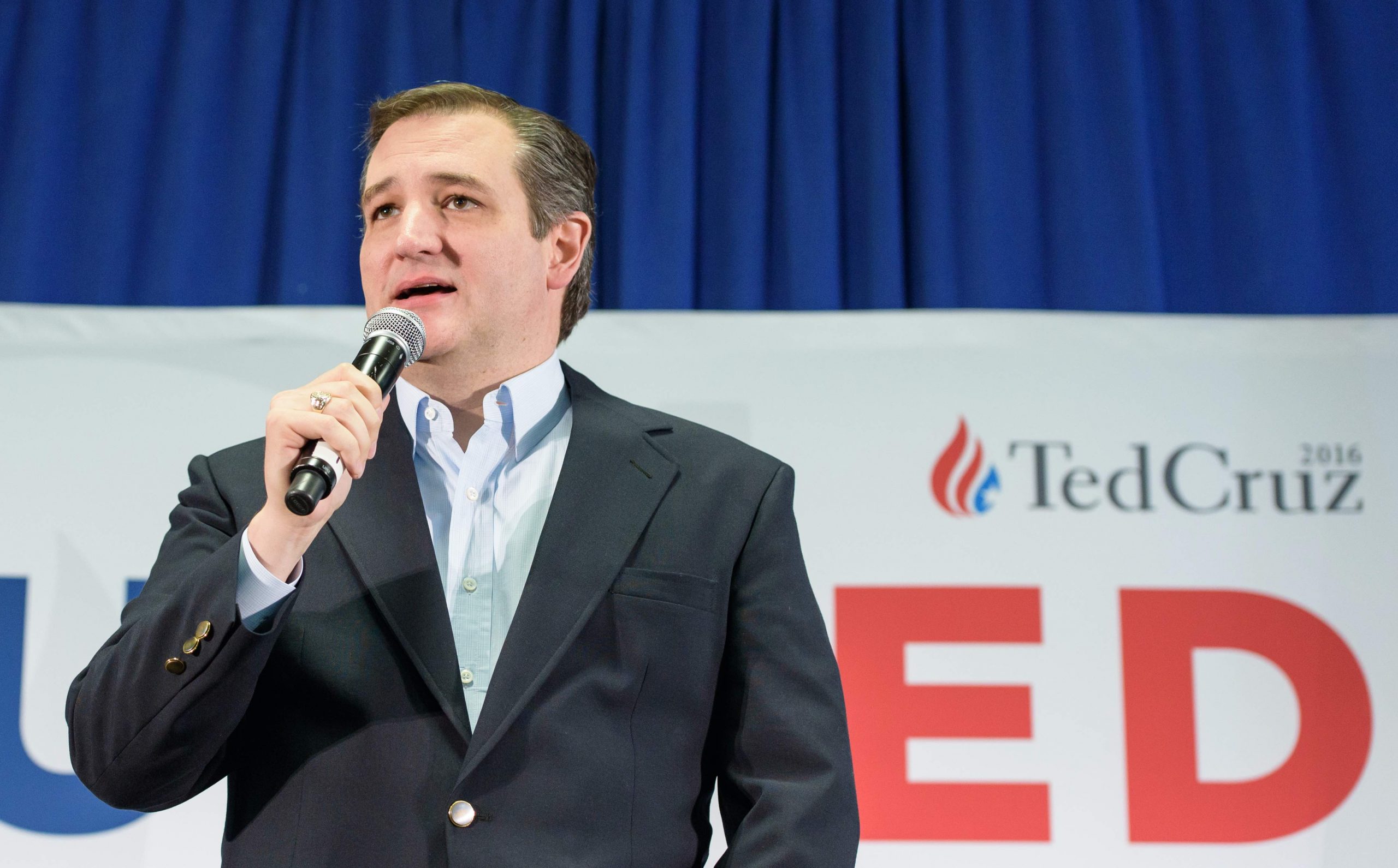 man in blazer speaks into handheld microphone in front of campaign sign