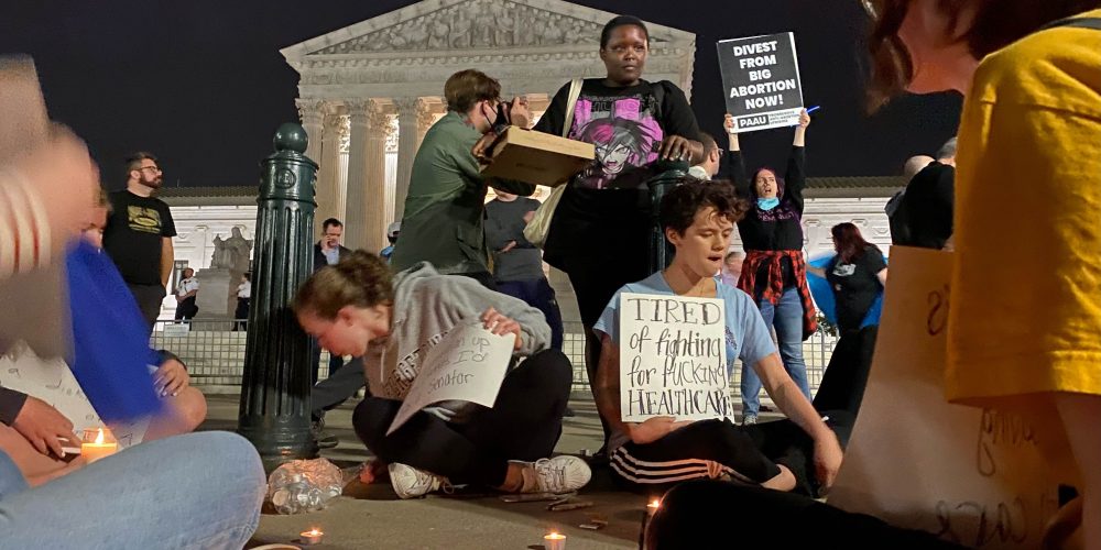 nighttime scene of people seated on ground with candles in front of supreme court
