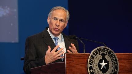 man holds both hands close to chest while speaking behind lectern bearing a texas seal