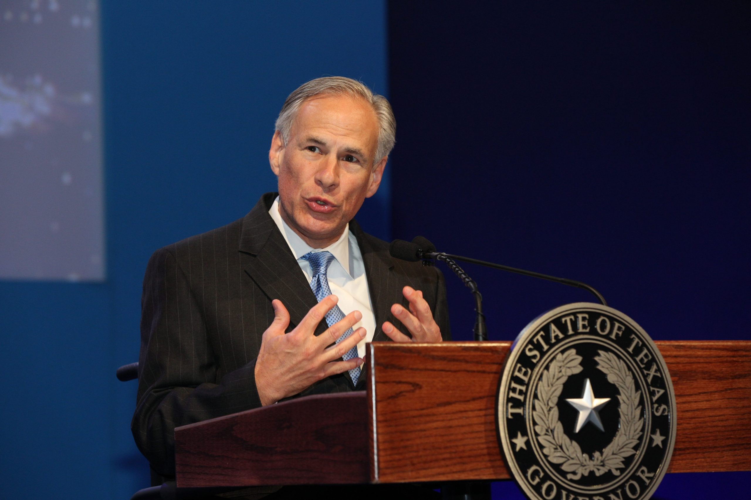 man holds both hands close to chest while speaking behind lectern bearing a texas seal