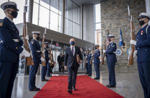 man wearing suit and face mask strides down red carpet as men in military garb stand at attention on both sides