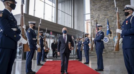 man wearing suit and face mask strides down red carpet as men in military garb stand at attention on both sides