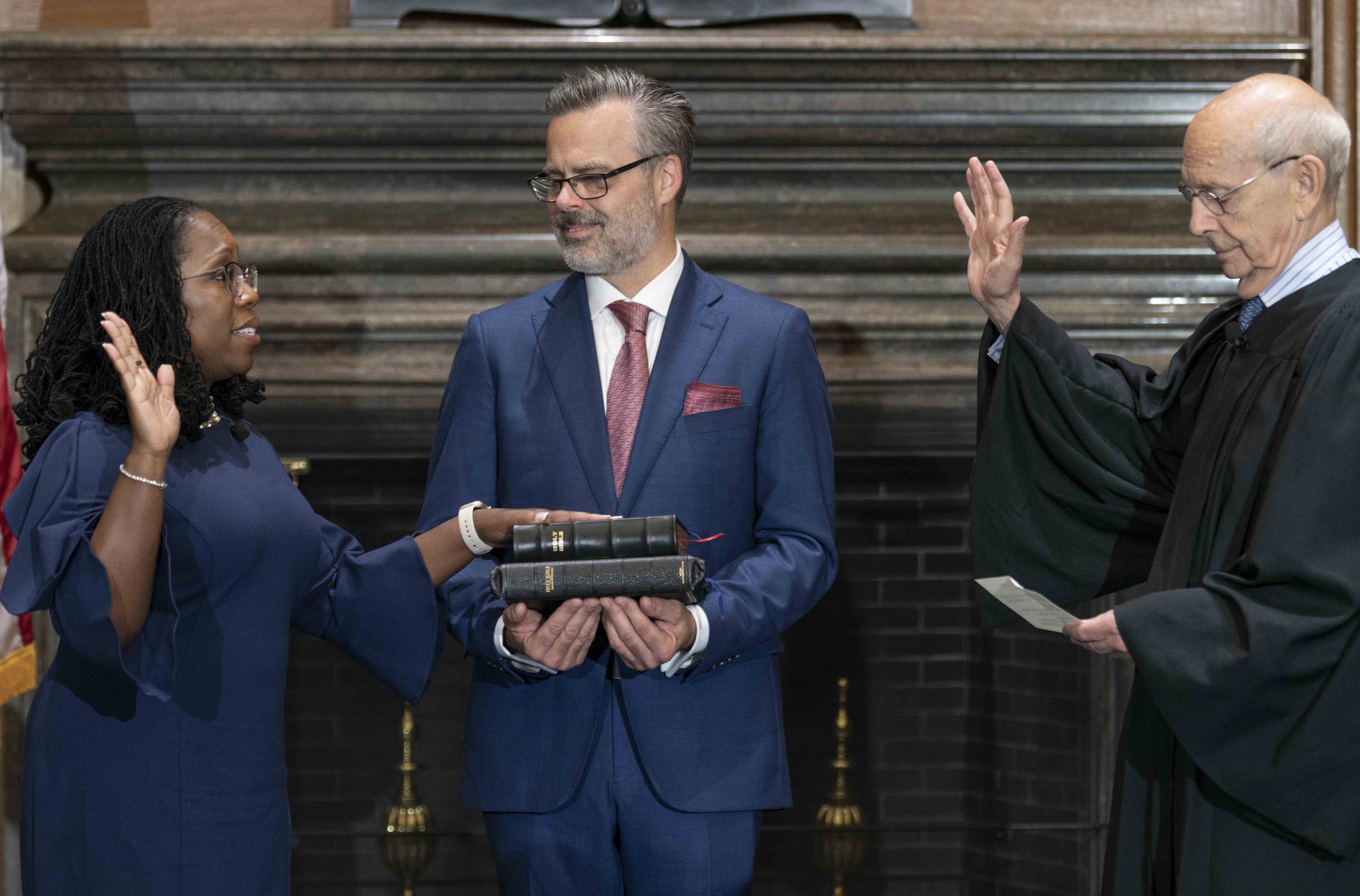 ketanji brown jackson stands with her right hand raised, stephen breyer stands facing her with his right hand raised while reading from piece of paper, jackson's husband stands between them looking at jackson