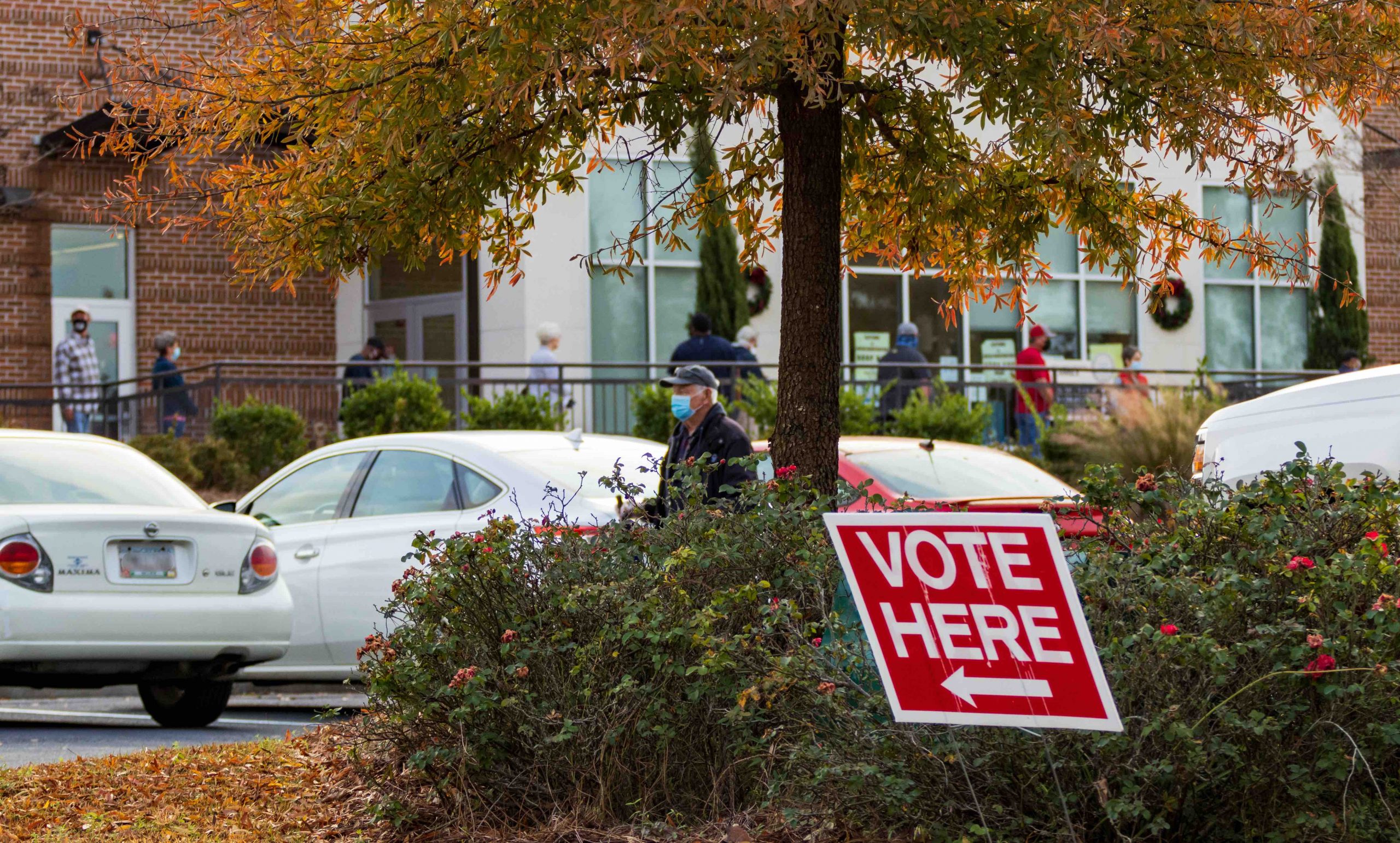 parking lot with "vote here" sign and line of people waiting outside in background