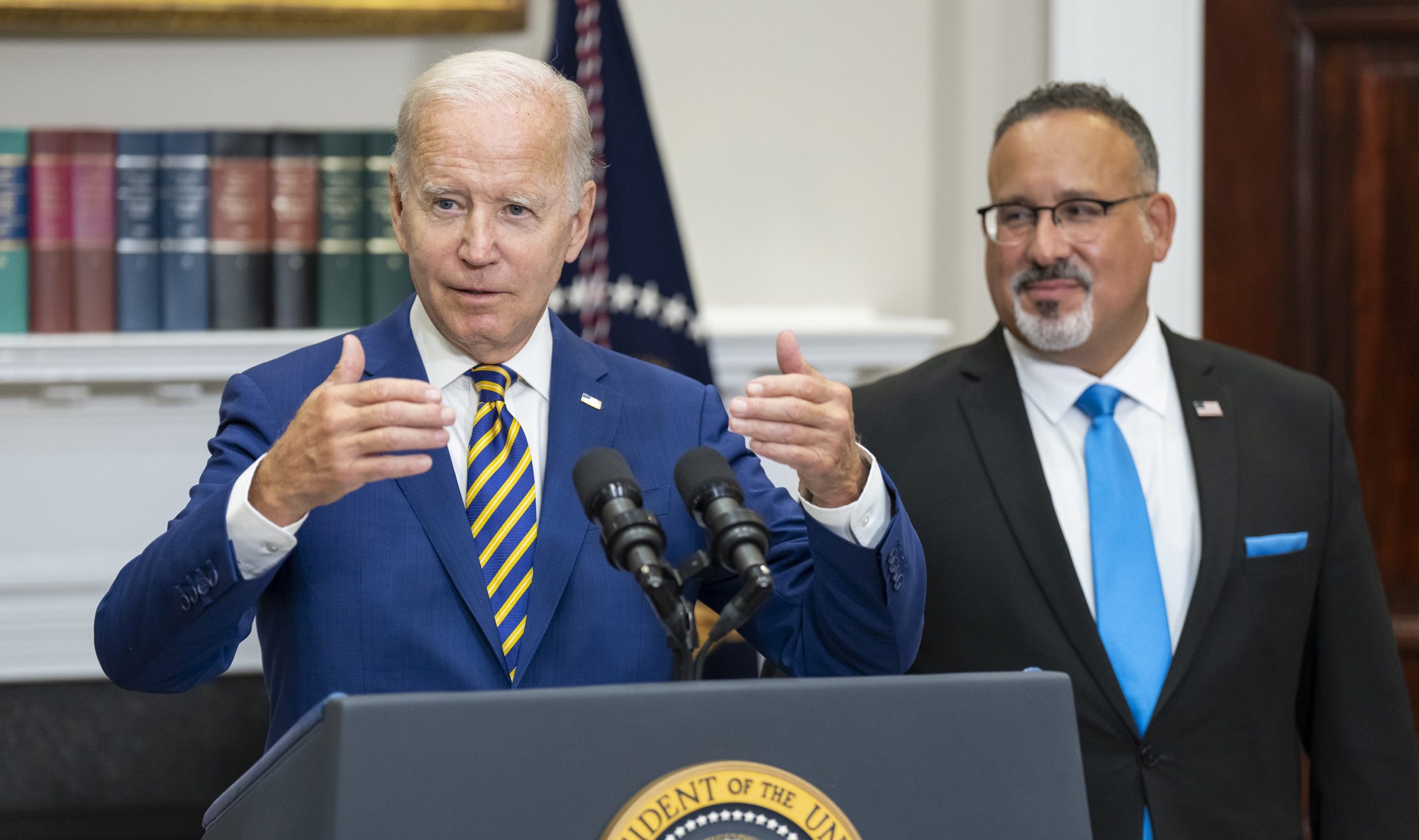 joe biden at lectern, gesturing with both hands, as miguel cardona looks on