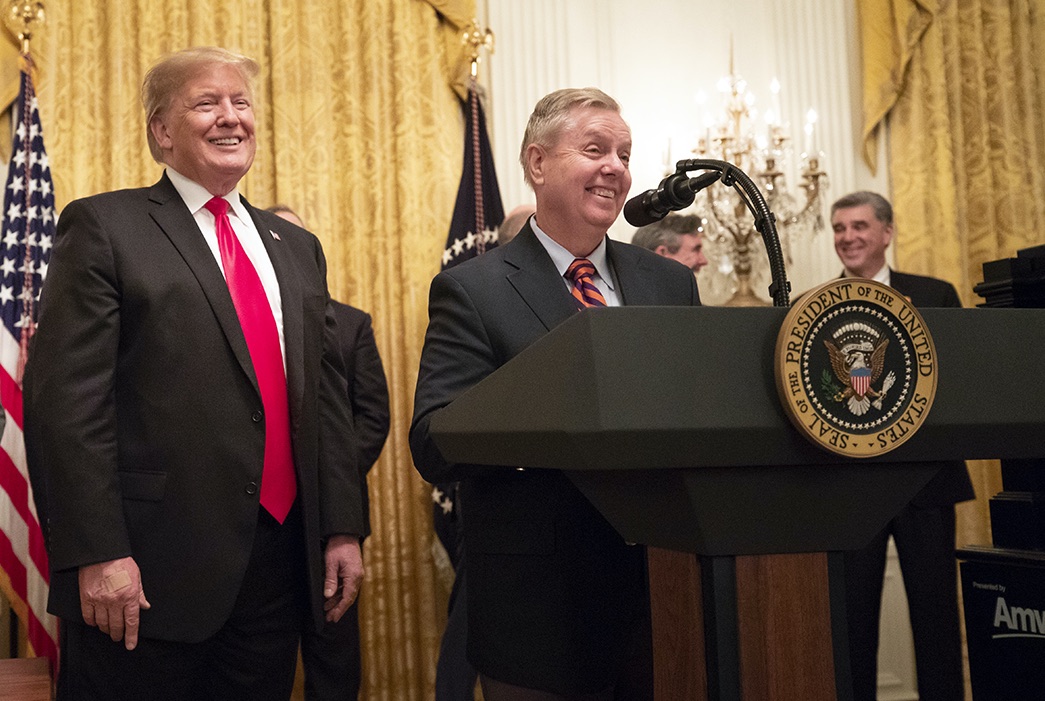 lindsey graham smiles while standing at lectern bearing presidential seal. donald trump stands alongside.