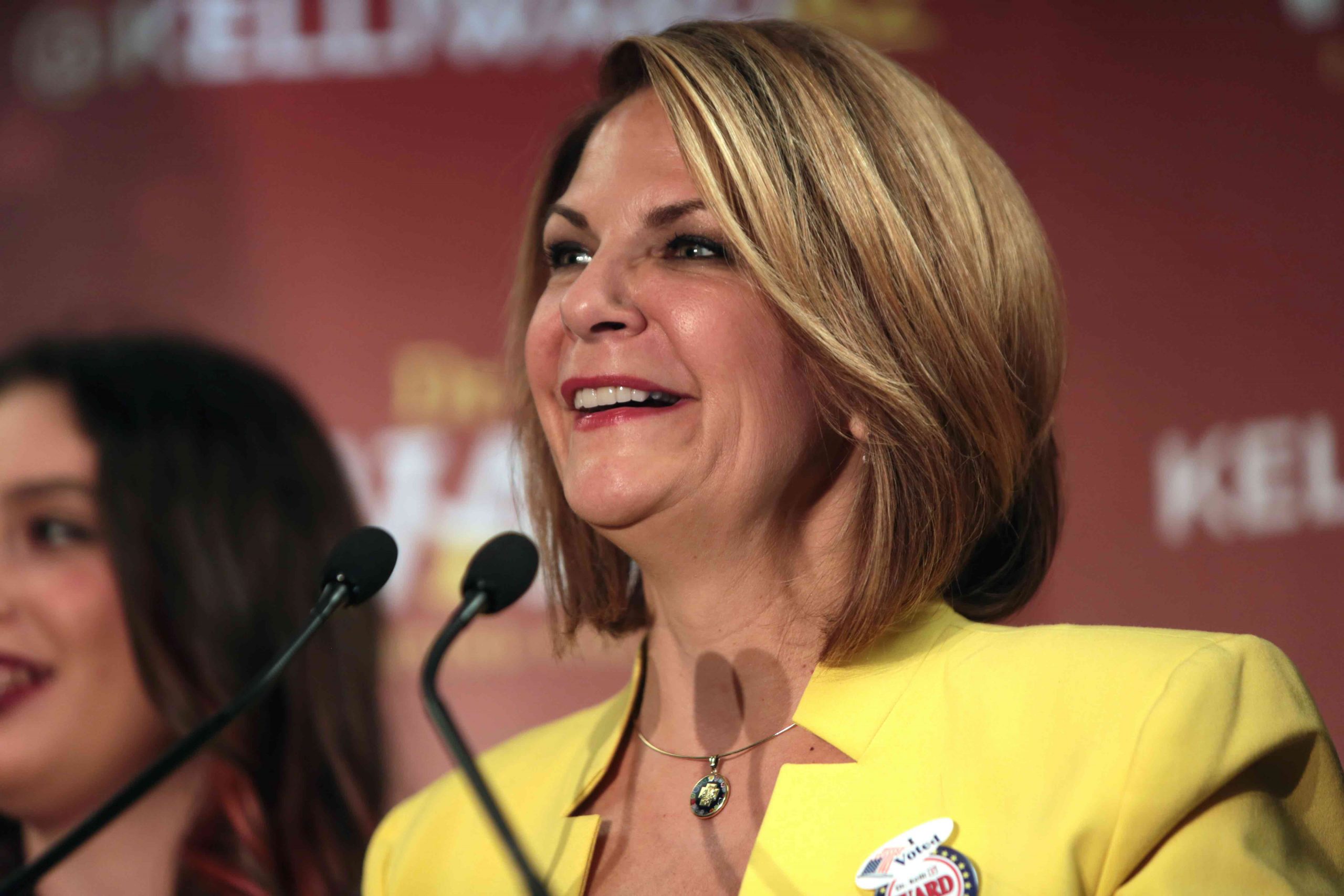 woman in yellow suit stands in front of two microphones