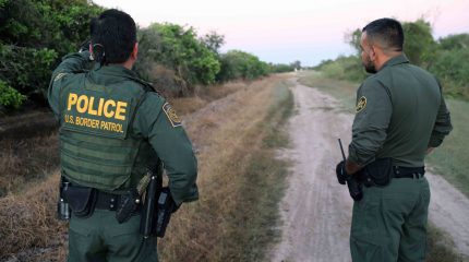 two men wearing border parol uniforms stand along dirt road with back to the camera. one points into the distance.