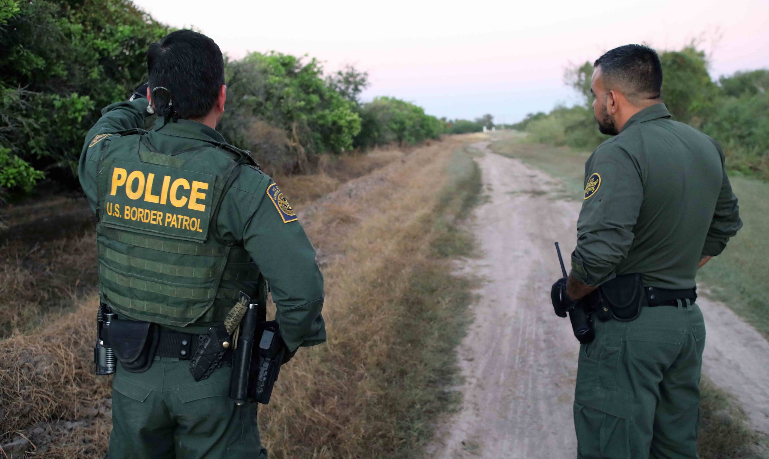 two men wearing border parol uniforms stand along dirt road with back to the camera. one points into the distance.