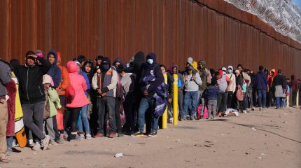 People wearing coats line up alongside a tall wall topped by razor wire.