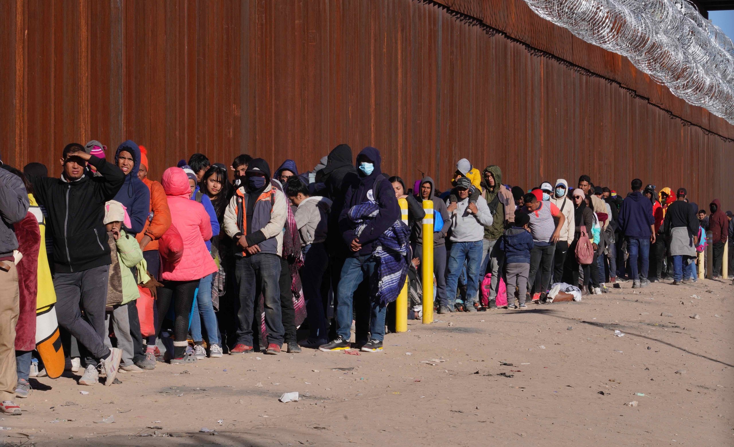 People wearing coats line up alongside a tall wall topped by razor wire.