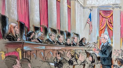 A man speaks at a lecture before nine justices on the bench who all seem to be asking him a question.