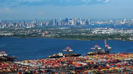 aerial view of busy seaport crowded with containers. a body of water separates the port in the foreground from the manhattan skyline in the background.