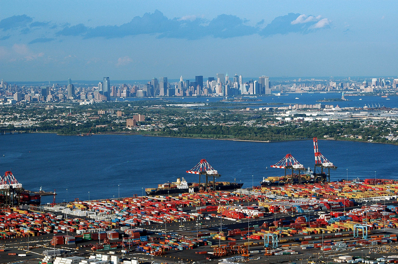 aerial view of busy seaport crowded with containers. a body of water separates the port in the foreground from the manhattan skyline in the background.