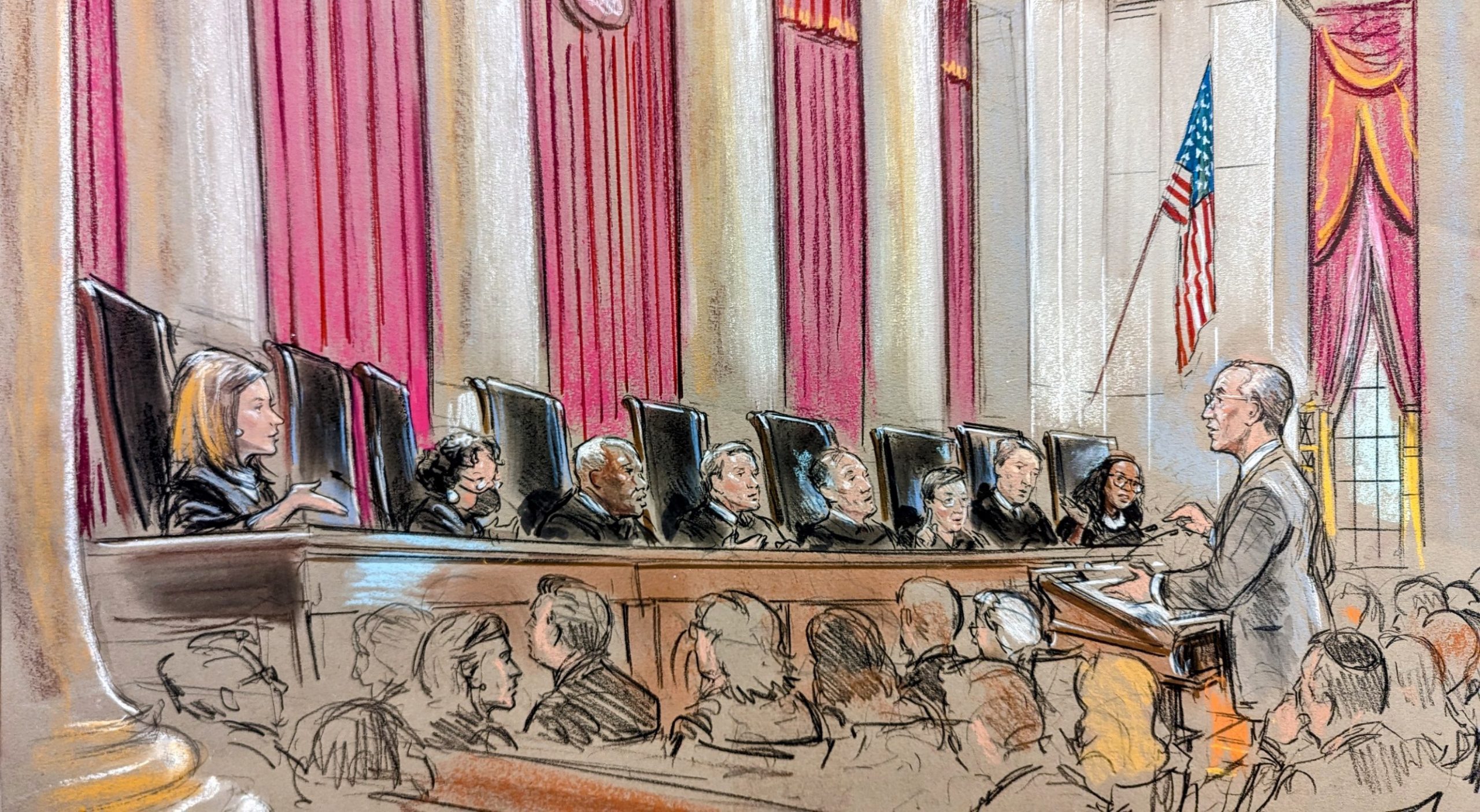 Eight justices on the bench speaking to and watching a man at the podium in a crowd of people.