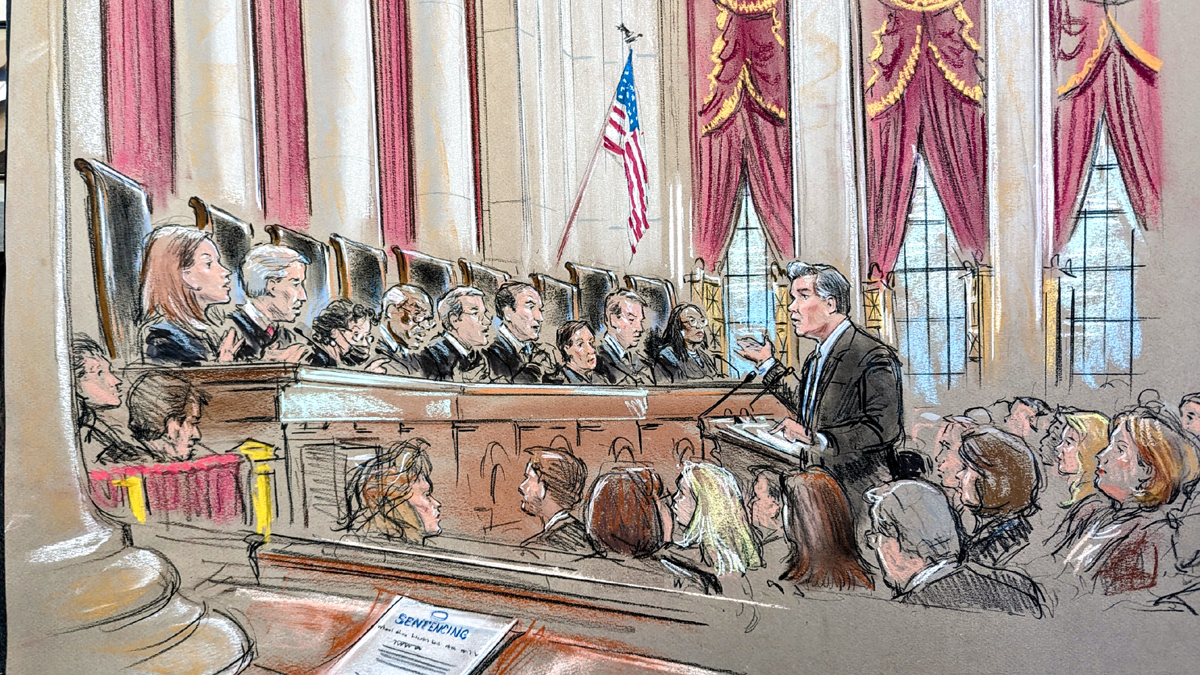 A wide shot of the court room. A man speaks from the lectern surrounded by a crowd as the justices speak from the bench.