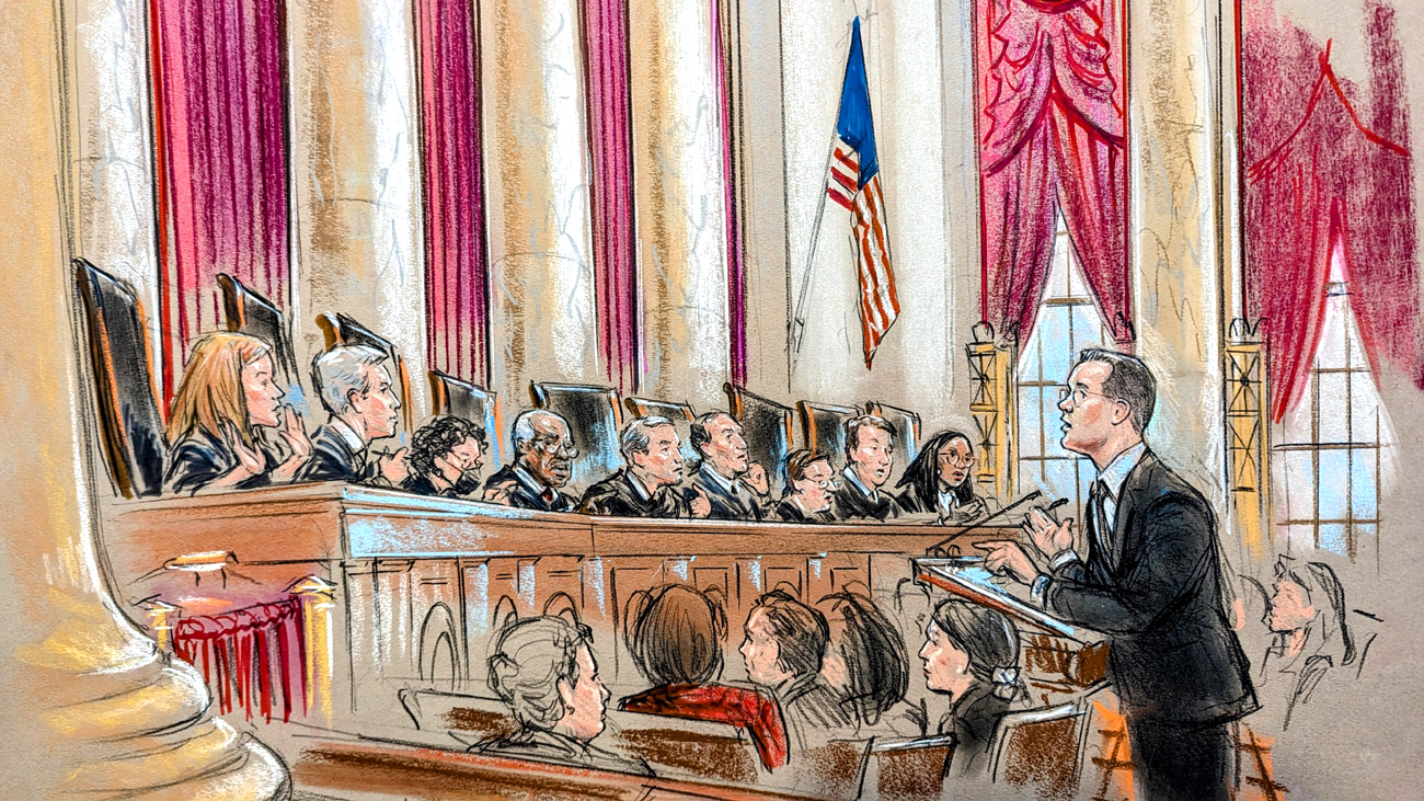 A man speaks to the full bench of justices from the lectern. A seated crowd listens.