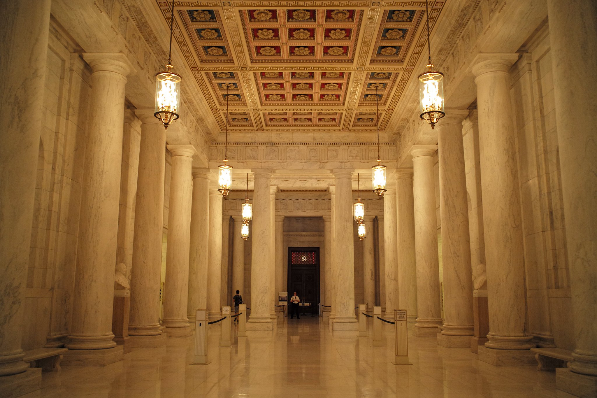 A long marble lobby with hanging lamps and marble columns. A guard waiting by a door at the end of the hall.