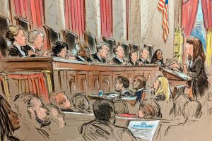 A woman at a podium speaks to a bench of justices as crowd looks on. 